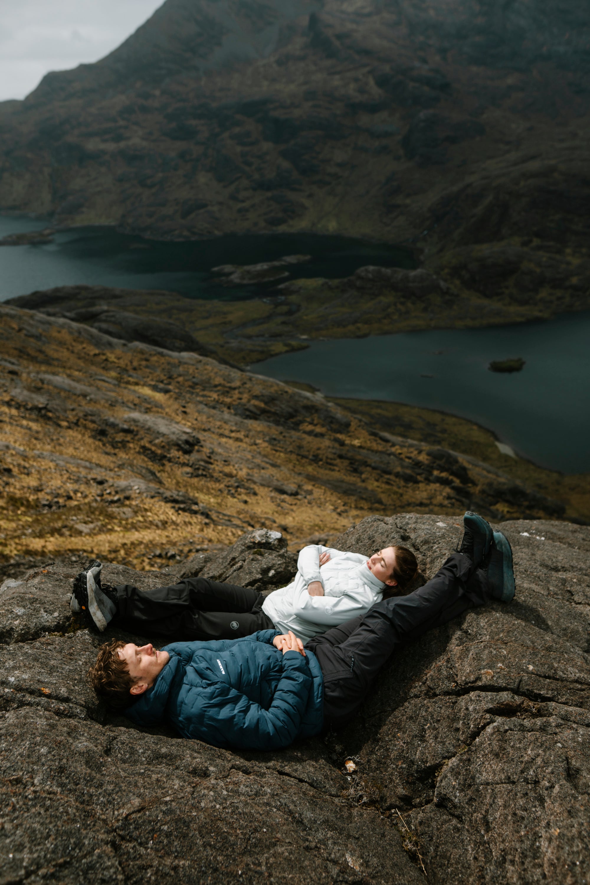 Two people in Mammut outdoor gear relax on lakeside rocks, with scenic mountains in the background, enjoying nature.